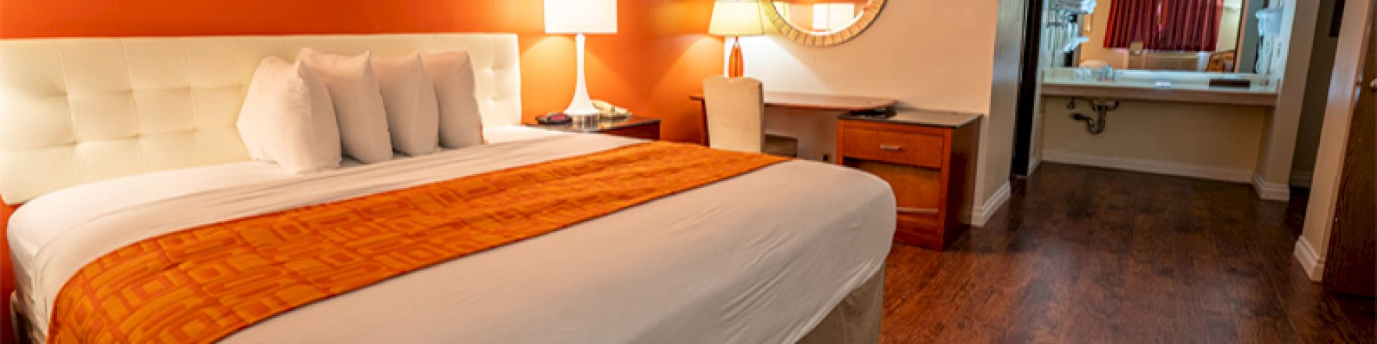 A cozy hotel room with a large bed, orange accent wall, lamps, desk, mirror, and a view into the bathroom area with a sink and mirror.