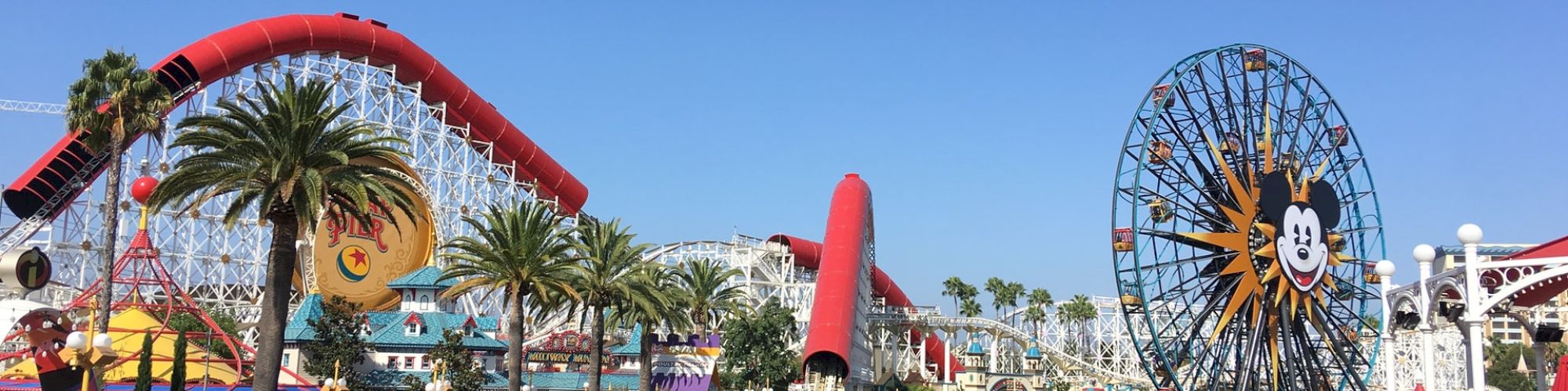 The image shows a theme park with a roller coaster, Ferris wheel with a cartoon character's face, palm trees, and water on a sunny day.