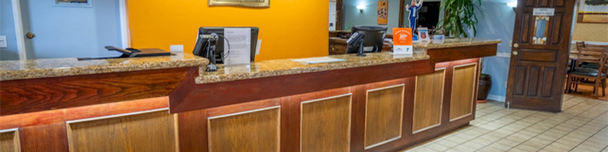 The image shows a hotel reception desk with wood accents, computers, informational materials, and a decorative plant in a warmly lit area.