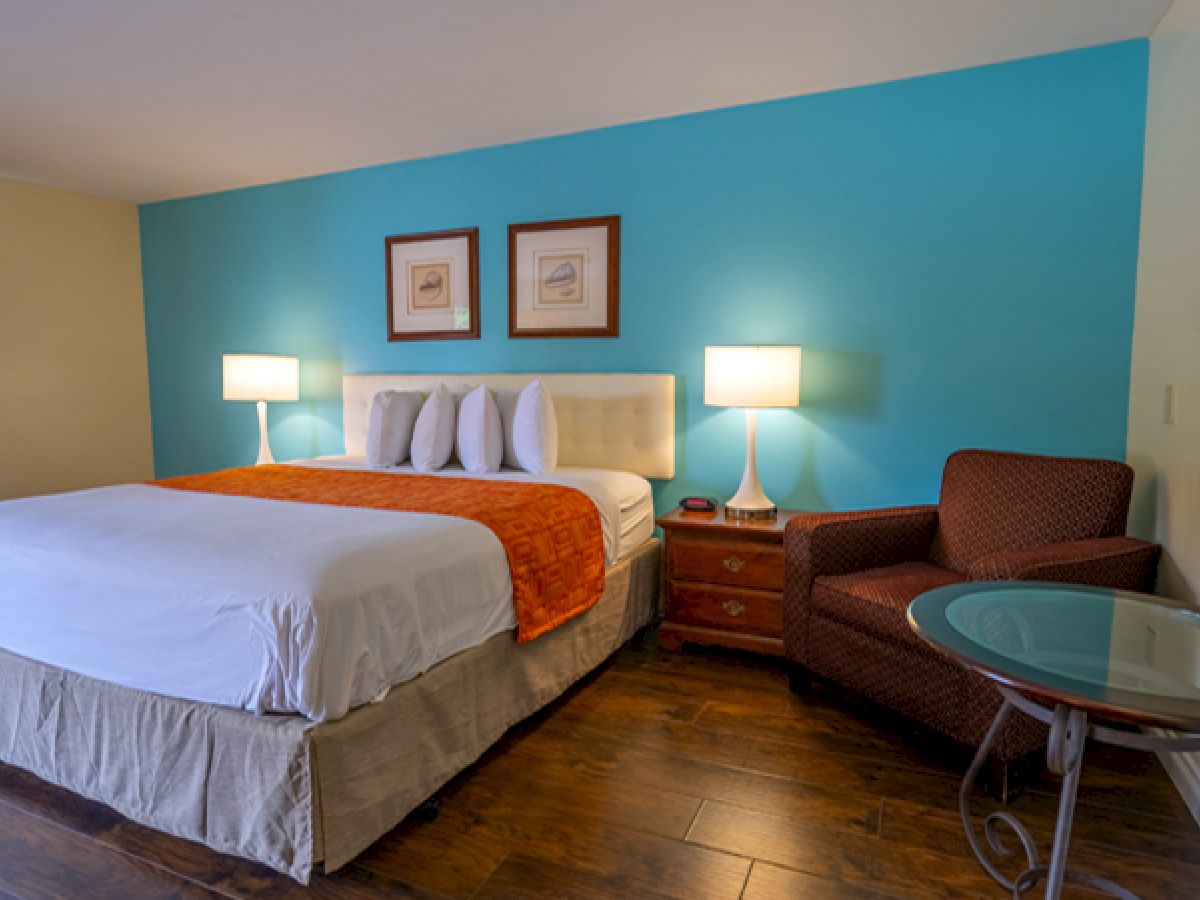 The image shows a hotel room with a bed, two lamps, a chair, a table, and a wall with blue paint, featuring a framed picture.