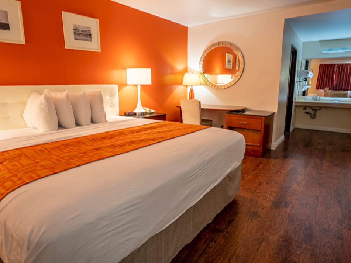 A hotel room featuring a large bed with orange accents, warm lighting, a desk, a mirror, and a visible bathroom area in the back.