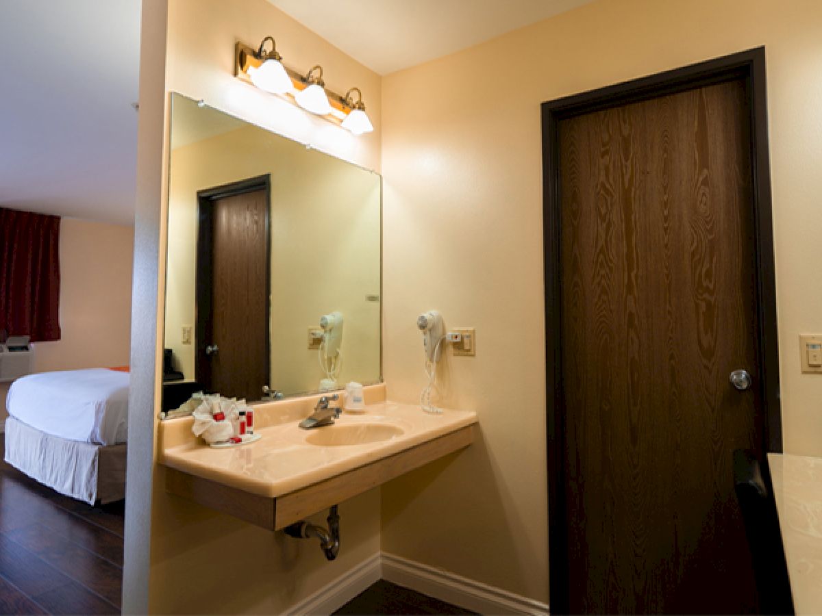 The image shows a hotel room with a bed, wood flooring, a vanity sink, mirror, and a door. Warm lighting and red curtains decorate the room.