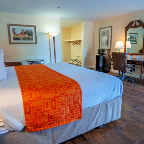 A hotel room with a bed, orange throw, TV, desk, and lamp. Walls have art, and flooring is wooden.