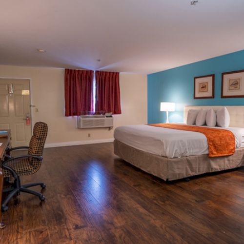 A well-lit hotel room with a bed, desk, chair, lamps, and wooden flooring. Walls are blue and white, with framed artwork.