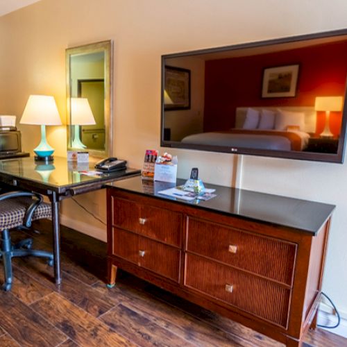 A hotel room desk with a chair, lamp, coffee maker, and a large TV on a cabinet. Hardwood floors and a mirror complete the setting.