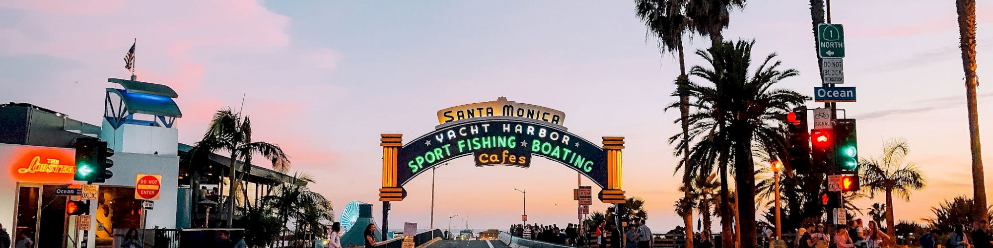 A crosswalk leads to a colorful archway reading "Sport Fishing Boating Cafes," with palm trees and bright skies in the background.