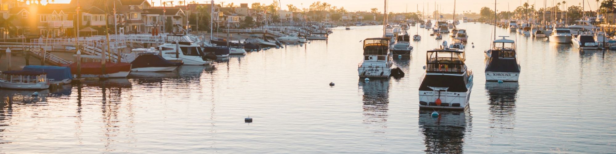 A serene harbor scene at sunset, featuring multiple docked boats, calm water, and a coastline lined with palm trees and buildings.