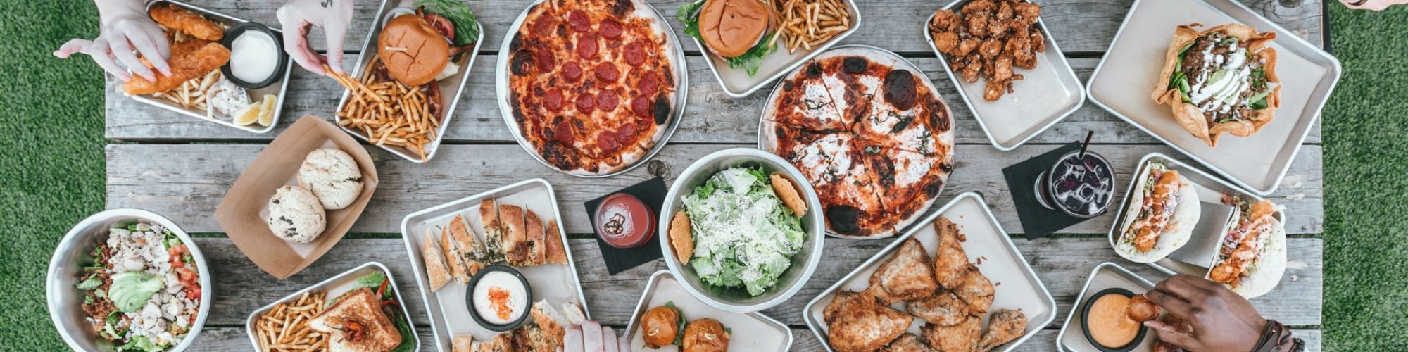 A group enjoys various foods on a picnic table, including pizza, wings, burgers, fries, and salad, with drinks placed around.